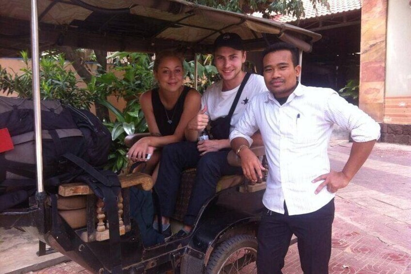 Three people on a wooden cart with a canopy, parked in an outdoor setting with plants and a building in the background. 2