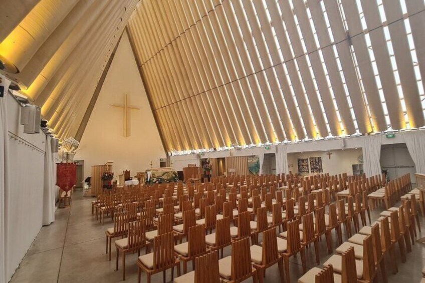 Ein Kircheninneres mit Holzbänken, einem Kruzifix an der Wand und einem großen Fenster. 2