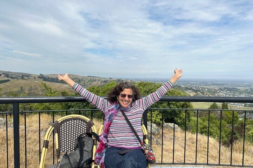 A person sitting on a chair with arms raised, overlooking a landscape with hills and a distant town. 2