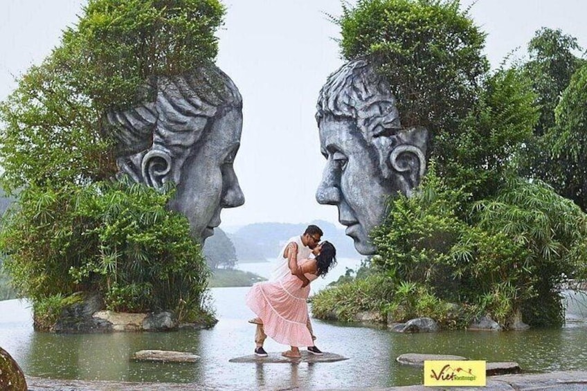 A couple embracing in a garden with large head sculptures and a body of water. 4