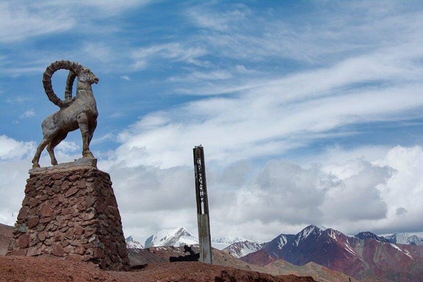 A stone ram statue on a rocky pedestal, a tall vertical structure, and a mountain range under a partly cloudy sky. 4