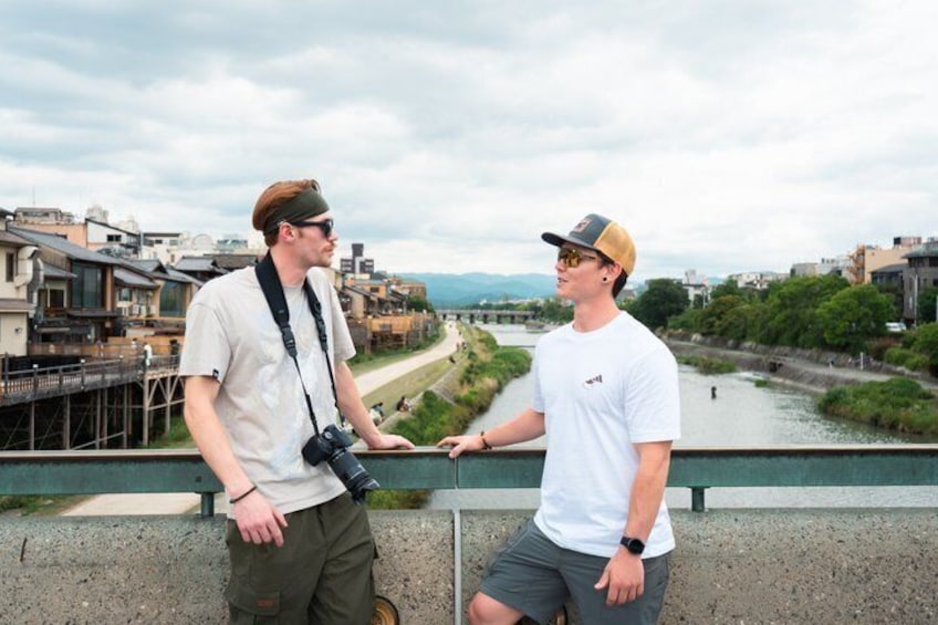 Dos personas de pie sobre un puente, con un río y edificios al fondo. 3