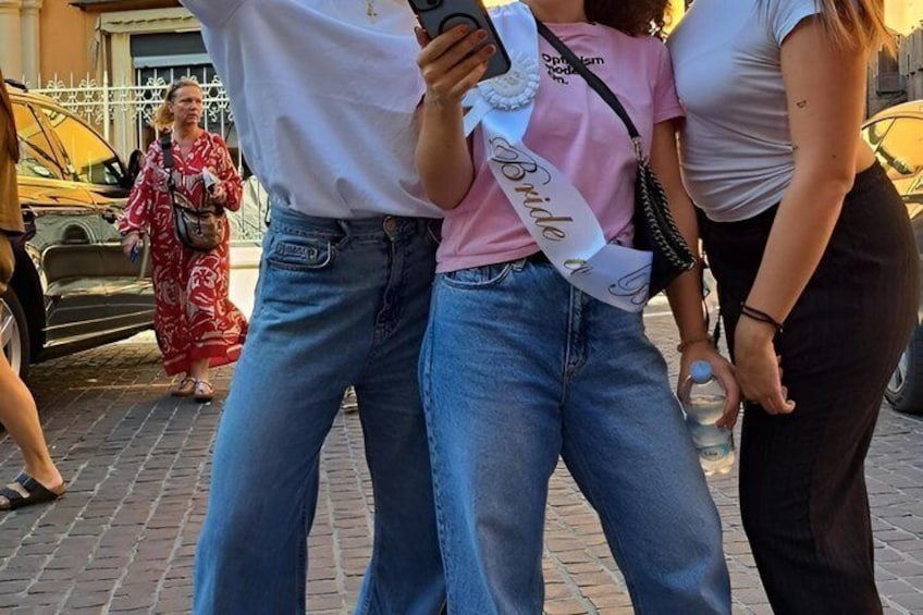 A person holding a smartphone, another person wearing a sash, and a car parked on the street. 3