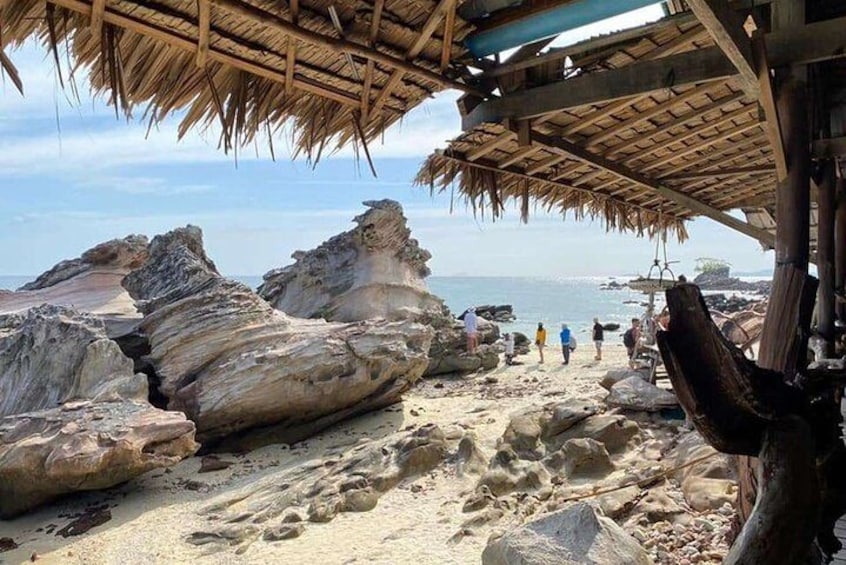 A rocky beach with large boulders, a thatched-roof structure, and people walking. 5