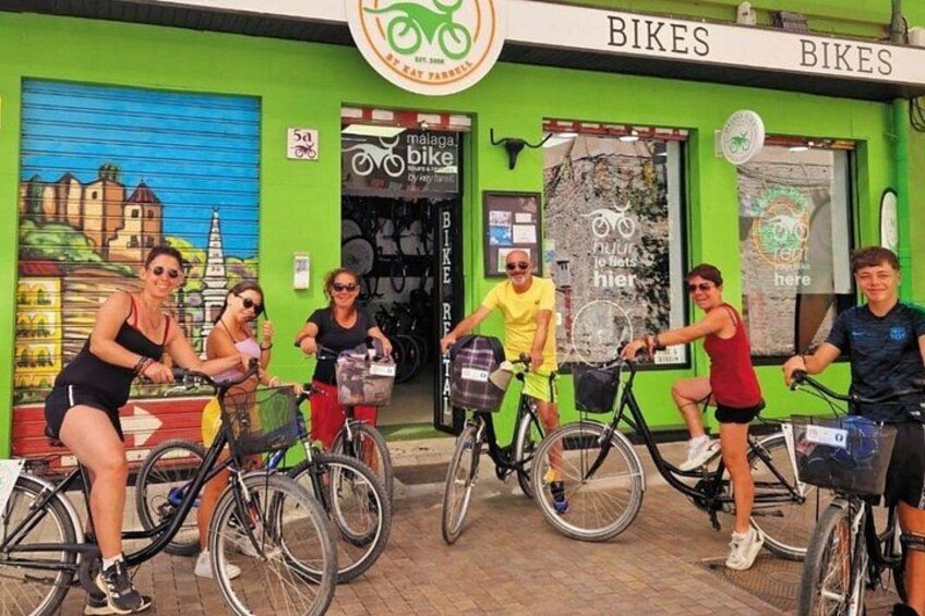 A group of people riding bicycles in front of a bike shop with a green facade and a mural of a castle. 2
