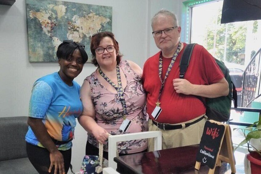 Three people standing indoors with a world map on the wall, a coffee table, and a sign that reads 'wifi coffee lounge'. 4