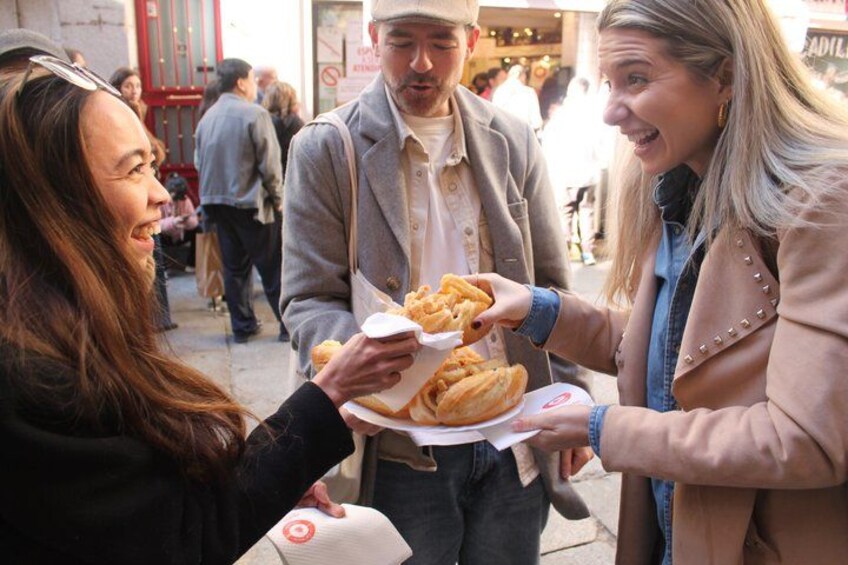 Tres personas sosteniendo comida, probablemente comida callejera, en un entorno urbano al aire libre con otras personas de fondo. 1