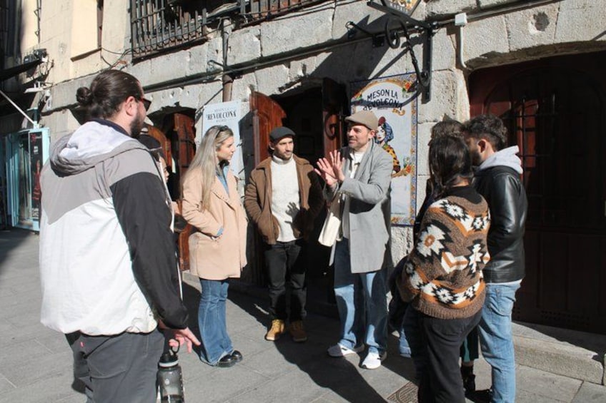 Un grupo de personas de pie en una calle, algunas sosteniendo bebidas, con la entrada de un edificio y un letrero al fondo. 3
