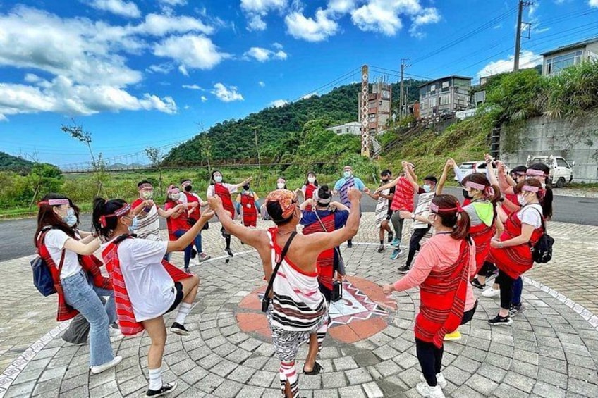 A group of people performing a synchronized dance on a paved area with a hilly landscape in the background. 1