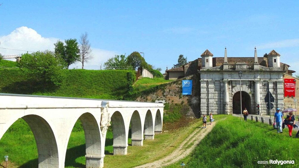 A stone bridge with arches, a historic building with a tower, and a grassy hill with trees. 2