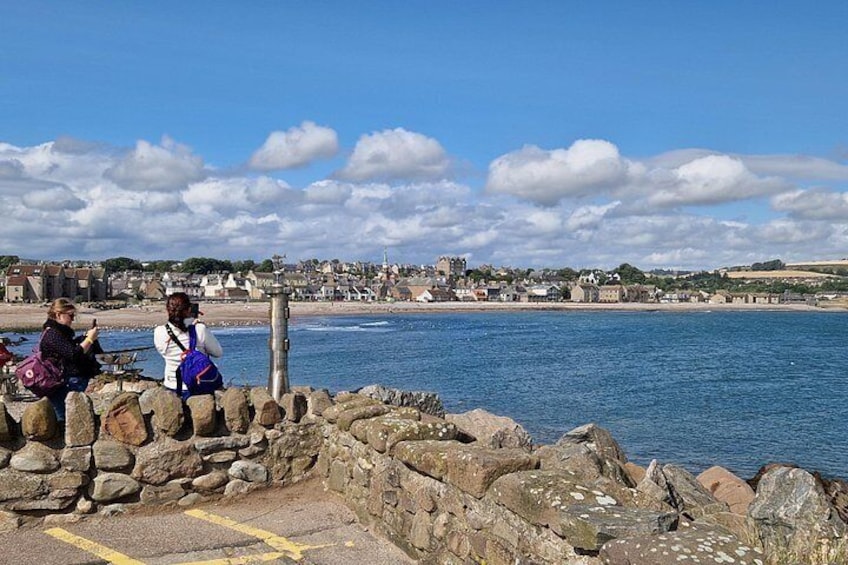 A coastal scene with a stone wall, people sitting, and a view of buildings and the sea. 3