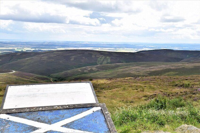 A sign on a grassy hill with a view of rolling hills and a cloudy sky. 5