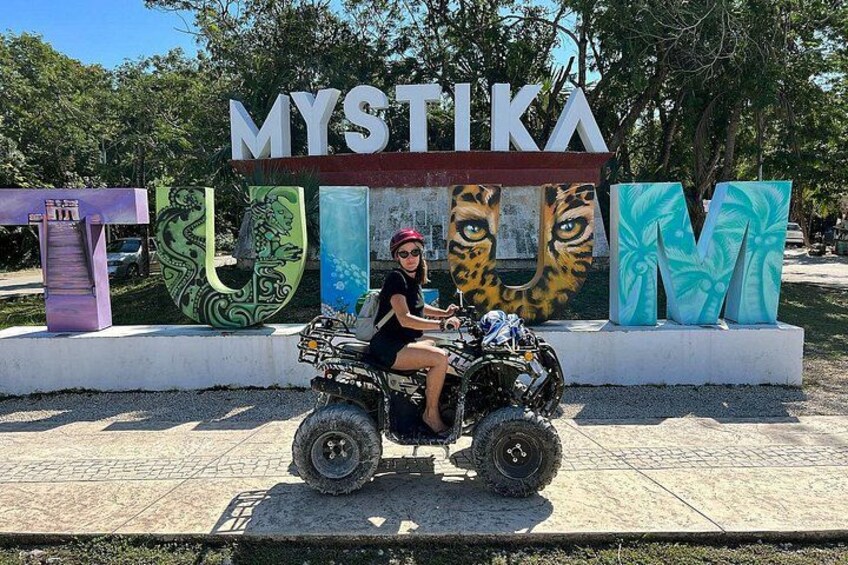 A person on a quad bike in front of a sign with large letters spelling 'MYSTIKA' and 'TUMI' with a tiger and palm tree design. 3