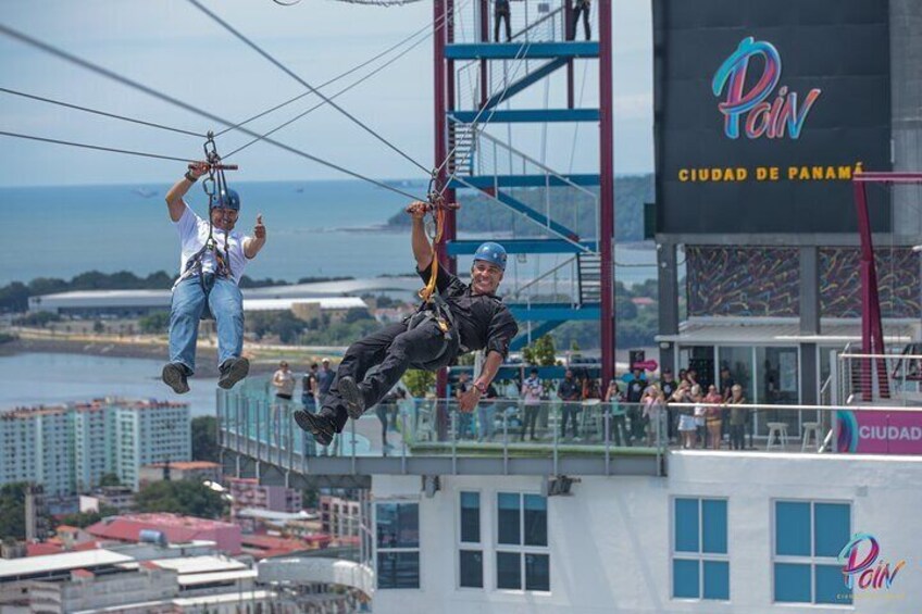 Två personer åker på en zipline över en stad med utsikt över havet och byggnaderna. 4