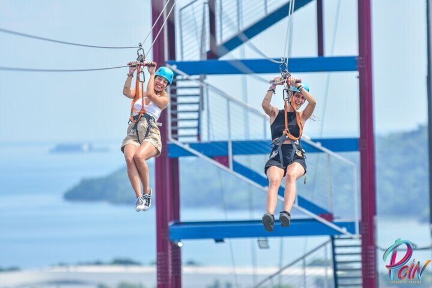 Två personer åker på en zipline över en bro, med en vacker utsikt över havet i bakgrunden. 3