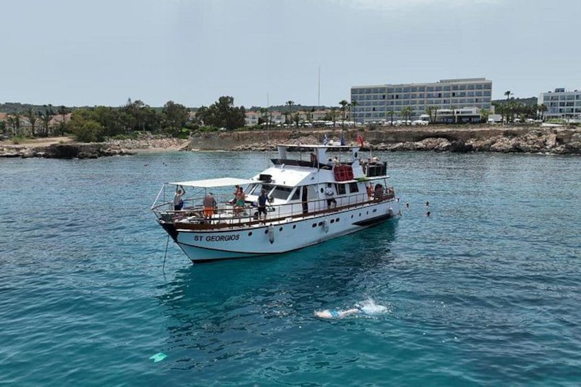 Ein weißes Boot namens ST GEORGIOS auf ruhigem Wasser mit einer felsigen Küstenlinie und Gebäuden im Hintergrund. 1