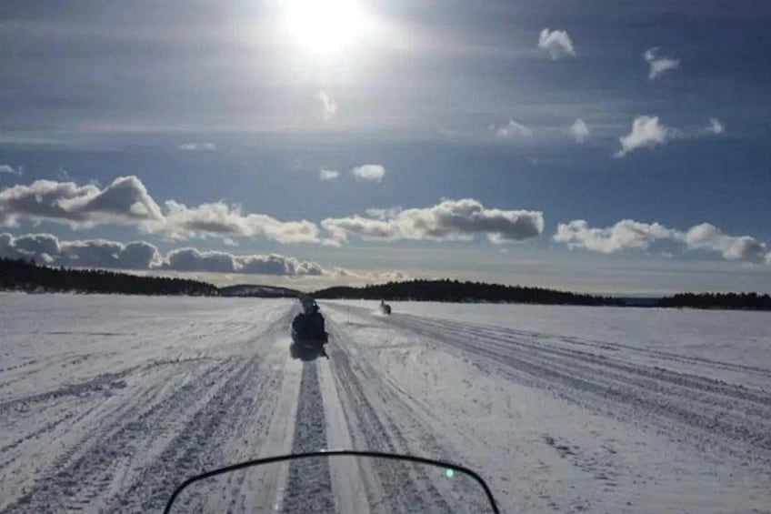 A snow-covered road with tire tracks, a clear sky, and distant trees. 1