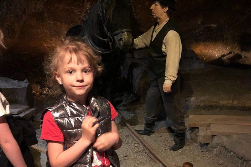 A person in a red shirt and a shiny vest, standing in a cave-like environment with rocky walls and a wooden structure. 2