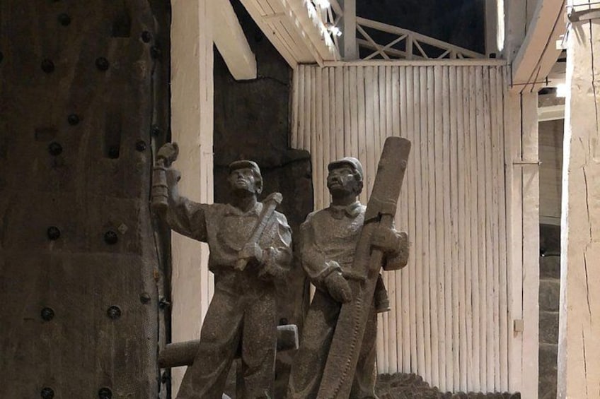 A statue of two figures, one holding a rifle and the other a pickaxe, in front of a wooden structure. 2