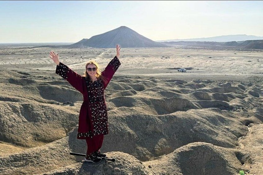 A person standing on rocky terrain with arms raised, a mountain in the background, and sparse vegetation. 2