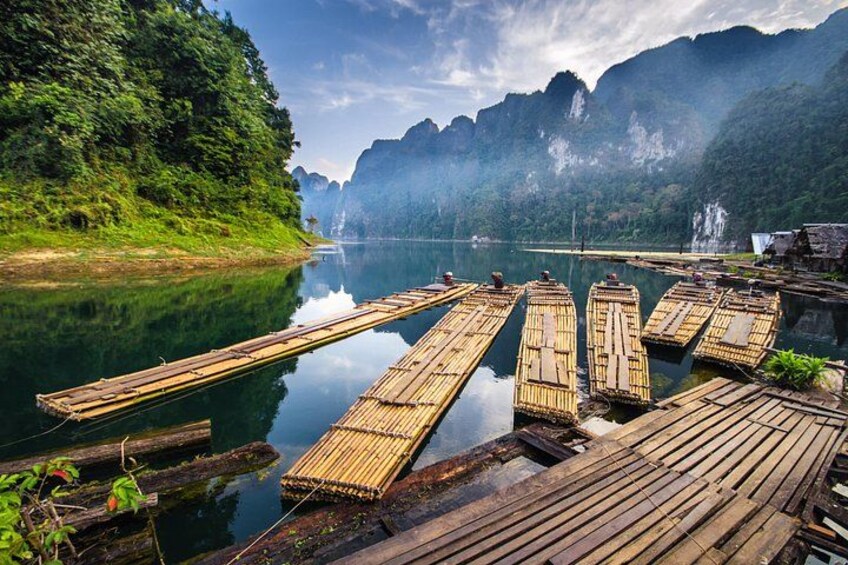 Une rivière paisible où des radeaux en bois sont amarrés le long de la rive, le tout entouré d’une végétation luxuriante et avec des montagnes en toile de fond. 2