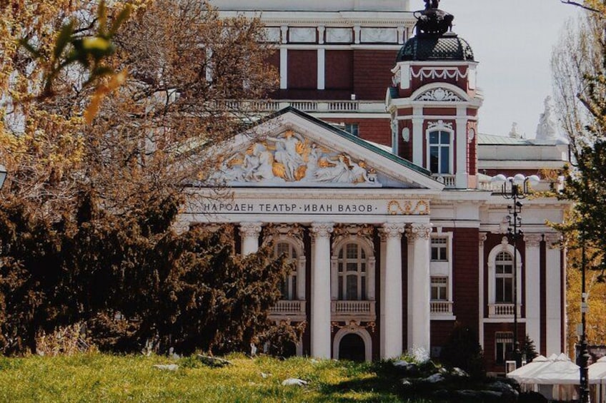 A historic theater building with classical architecture, surrounded by trees and a clear sky. 2