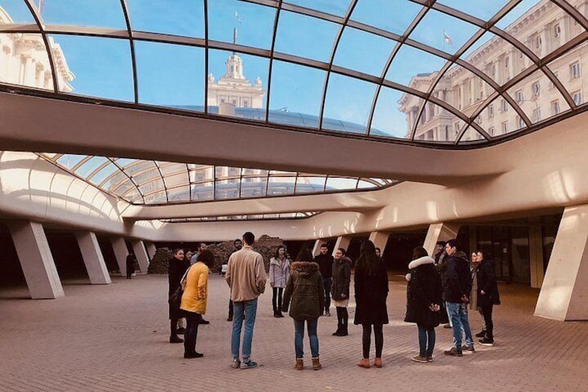 Una estructura arquitectónica moderna con techo de vidrio, personas adentro y edificios visibles a través del vidrio. 4