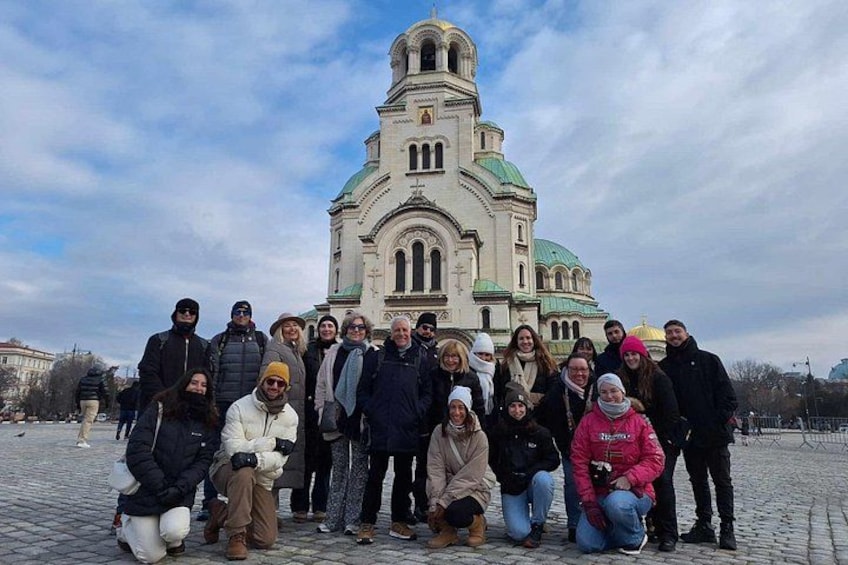 Een groep mensen poseert voor een grote kerk met een groene koepel en een hoge klokkentoren. 3