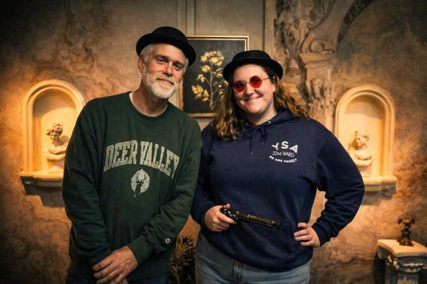 Two people standing indoors with a stone wall and sculptures in the background. 2