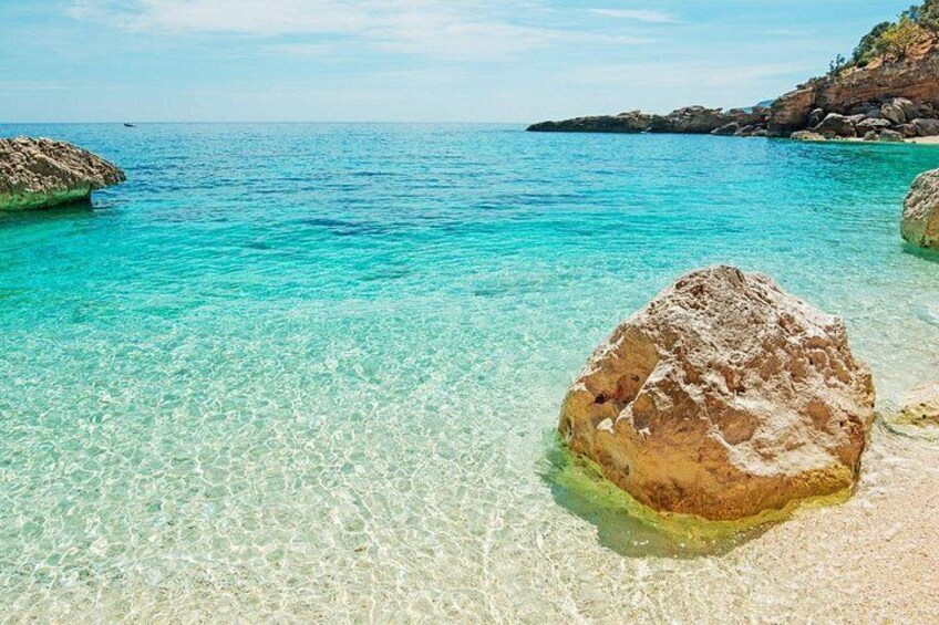 A clear turquoise sea, a large rock in shallow water, and rocky cliffs in the background. 2