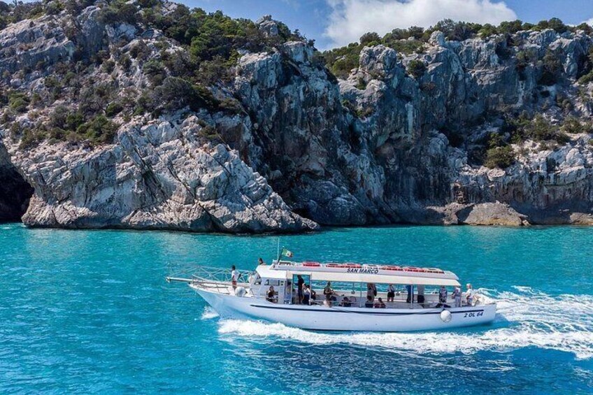 A boat named 'San Marco' on turquoise water with rocky cliffs in the background. 1