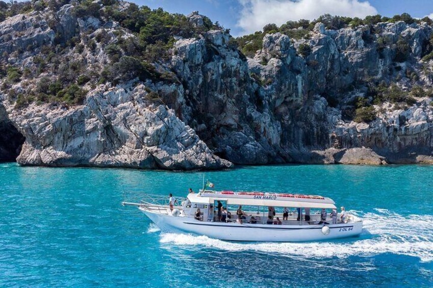A white boat named 'San Marco' with passengers, sailing on turquoise water with rocky cliffs in the background. 1