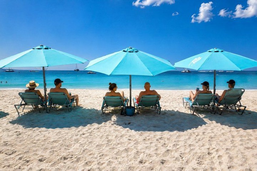A beach with people sitting under blue umbrellas, a clear sky, and the ocean in the background. 3