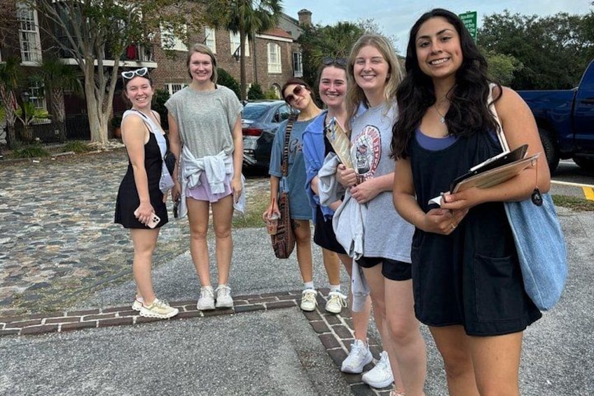 A group of people standing on a paved area with a brick sidewalk, parked cars, and a building in the background. 2