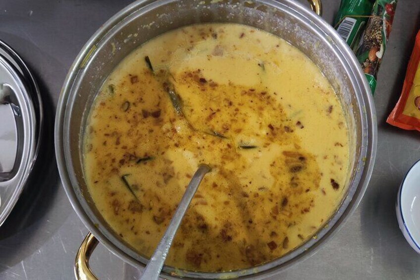 A pot of yellow curry with a spoon, spices, and a metal lid. 1