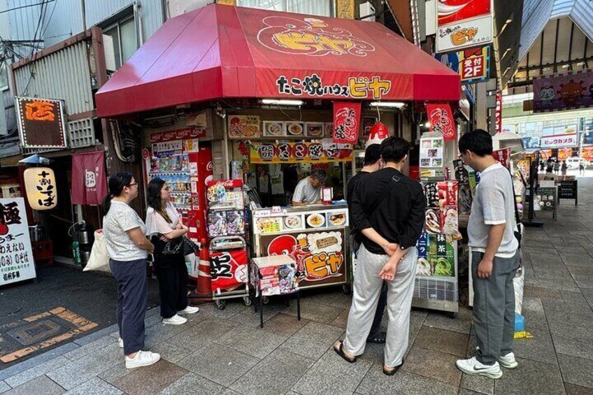 A street food stall with a red umbrella, surrounded by people. 4