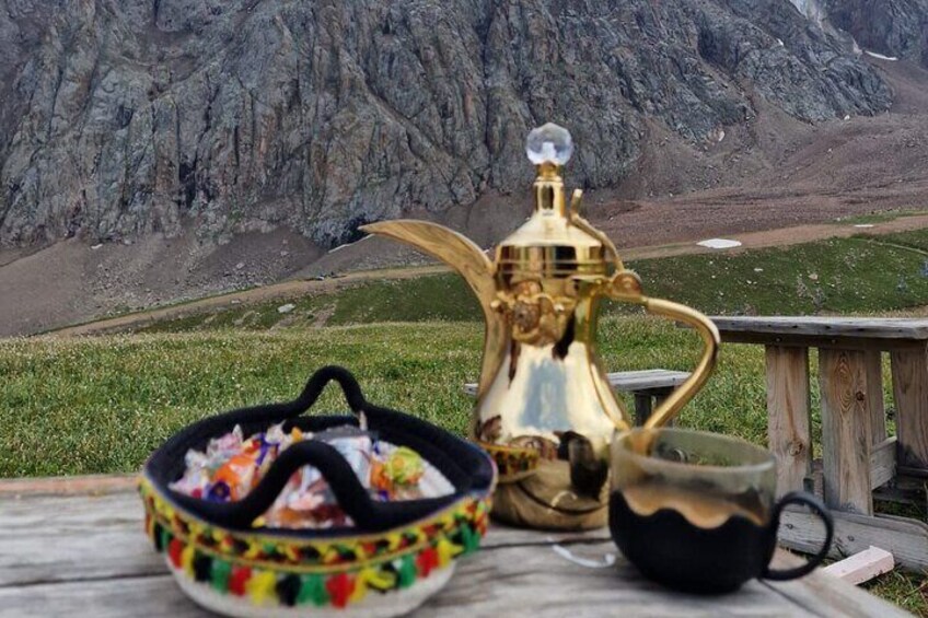 A brass teapot and a black cup on a wooden table with a colorful basket in the foreground, set against a mountainous landscape. 2