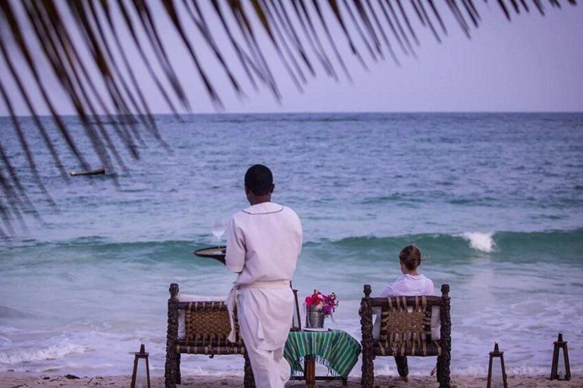 Two people sitting on a bench facing the ocean, with a table and chairs in the foreground. 3