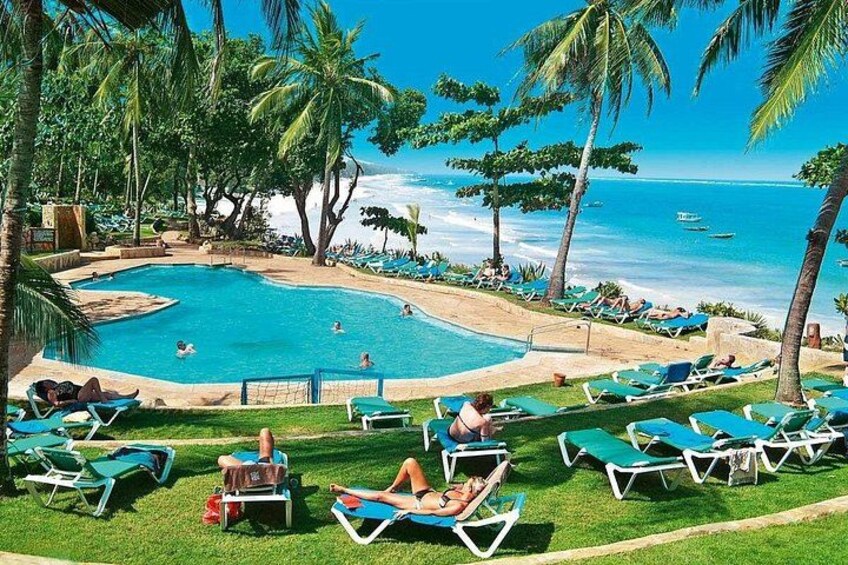 A pool with people swimming, surrounded by palm trees and beach chairs. 1
