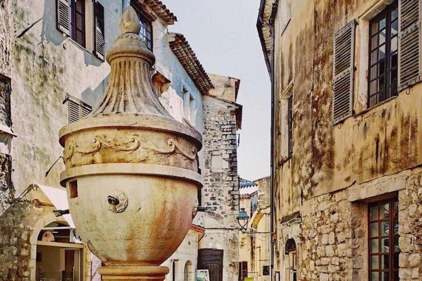 A narrow street with old buildings, a large ceramic pot, and a stone tower. 3