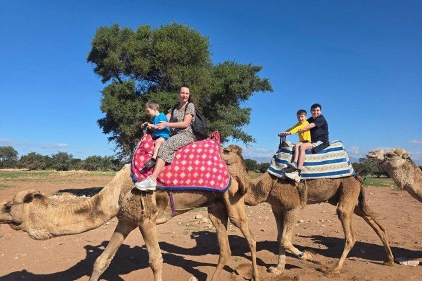 Tre persone cavalcano dromedari in un paesaggio desertico, con vegetazione rada e cielo sereno. 5