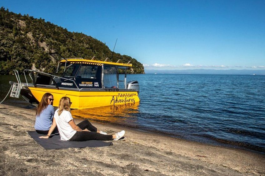 Two people sitting on a beach with a yellow boat named 'Tanjoa Adventures' in the background. 1