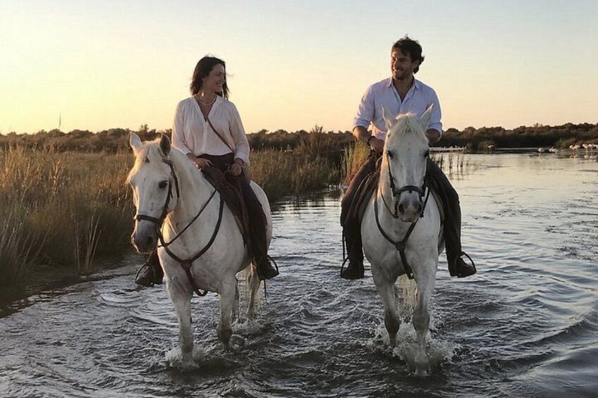 Two people riding horses through a shallow water body with tall grasses and a clear sky in the background. 1