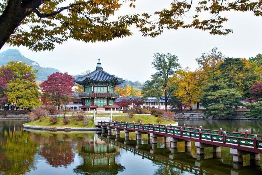 A traditional pavilion by a calm lake with autumn foliage and a wooden bridge. 1