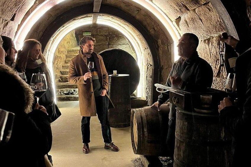 A group of people in a stone-walled tunnel, some holding wine glasses, and a man in a coat standing in the center. 2