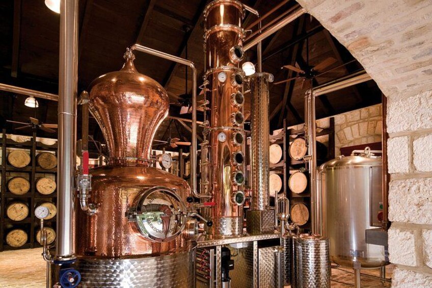 A copper distillation apparatus in a brewery setting with wooden barrels in the background. 2