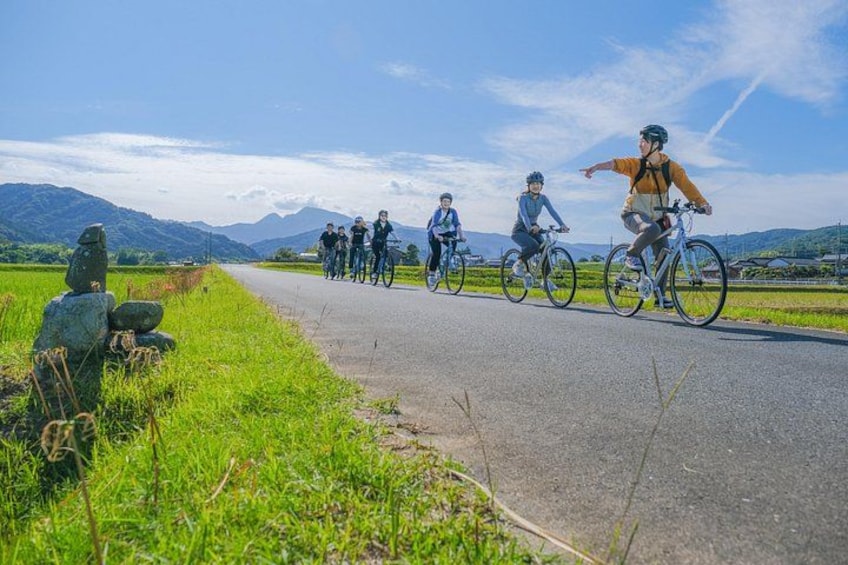A group of cyclists on a paved road with a rural landscape, including grass, a stone structure, and distant mountains. 1