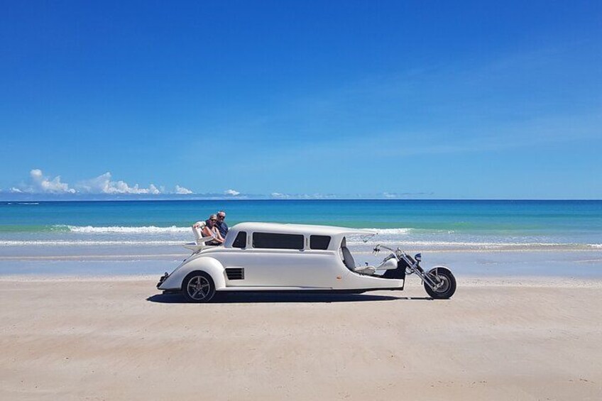 Een witte oldtimer geparkeerd op het strand, met twee personen achterin. 4