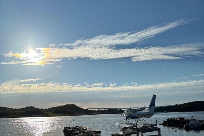 Ein Amphibienflugzeug auf dem Wasser, eine Anlegestelle und eine hügelige Landschaft unter klarem Himmel. 3