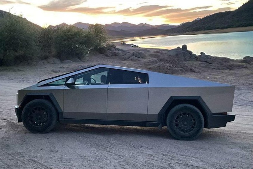A silver vehicle parked on a sandy beach with a lake and mountains in the background. 5